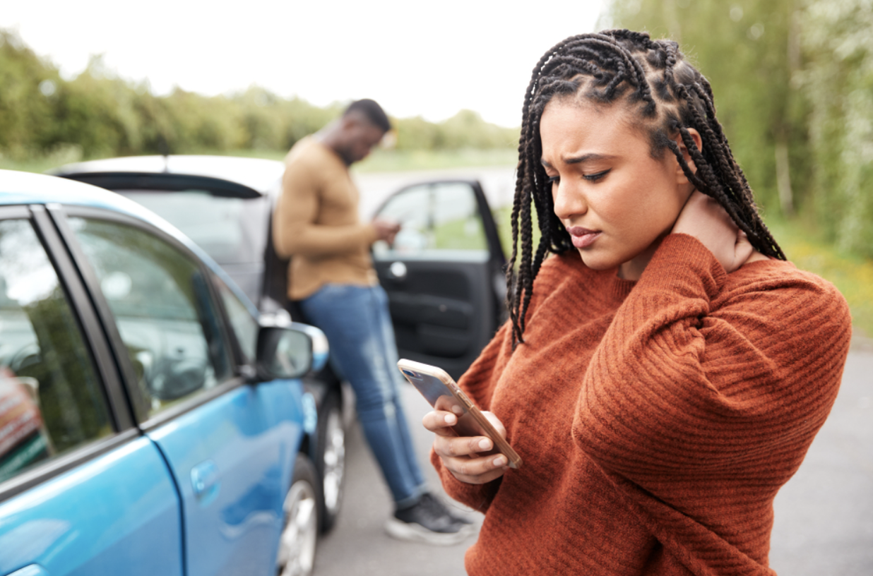 Woman by car, checking phone after a car accident, resulting in neck pain.