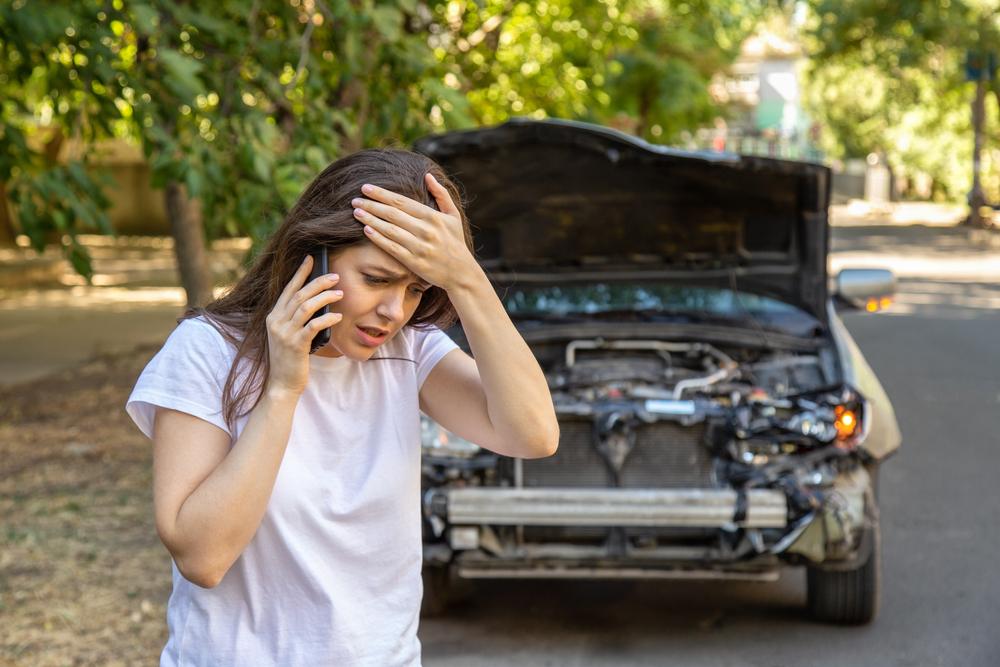 Driver woman portrait in front of wrecked car in car accident. Scared woman in stress holding her head after auto crash calling to auto insurance for help. Dangerous road traffic situation