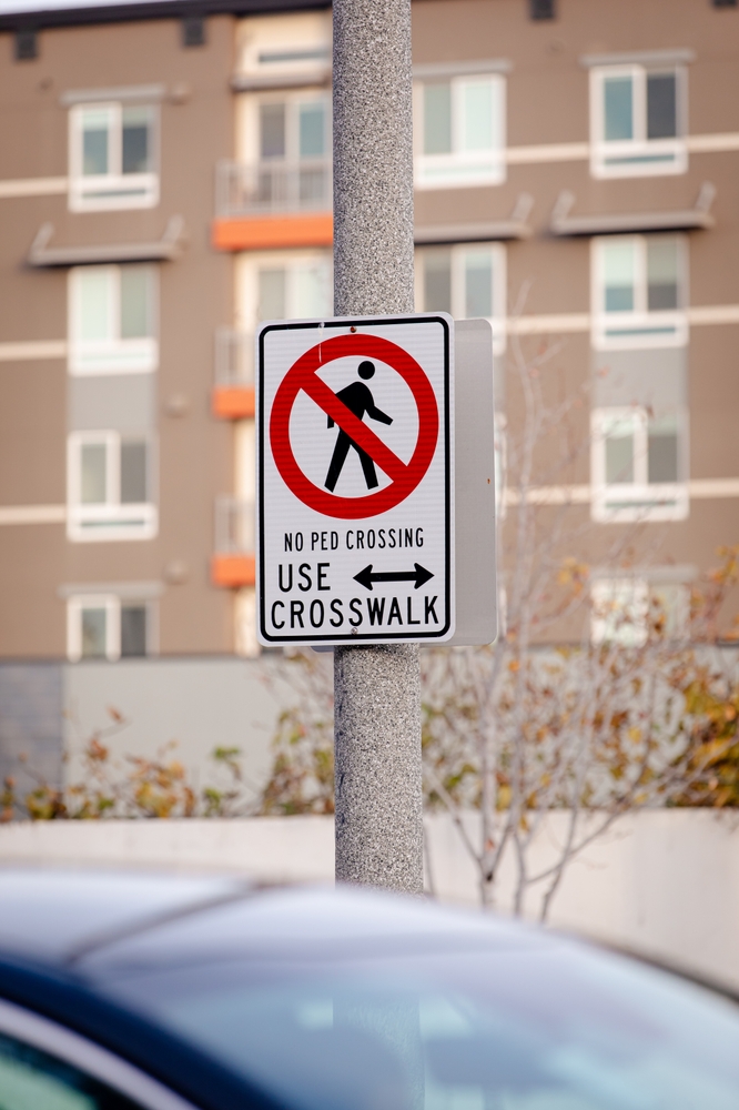 No Pedestrian Crossing Use crosswalk metal road sign on a lamp post o a street, blurred apartment building in the background