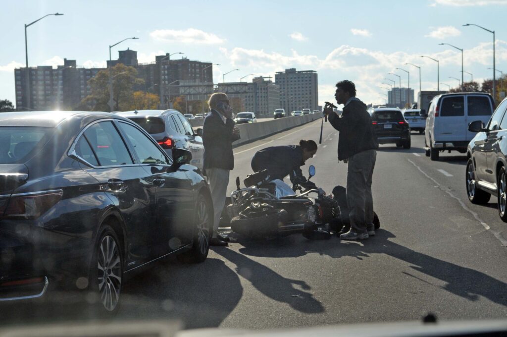 Bystanders and a motorcyclist gather around a downed bike on a busy highway following an accident to offer assistance and call for help.