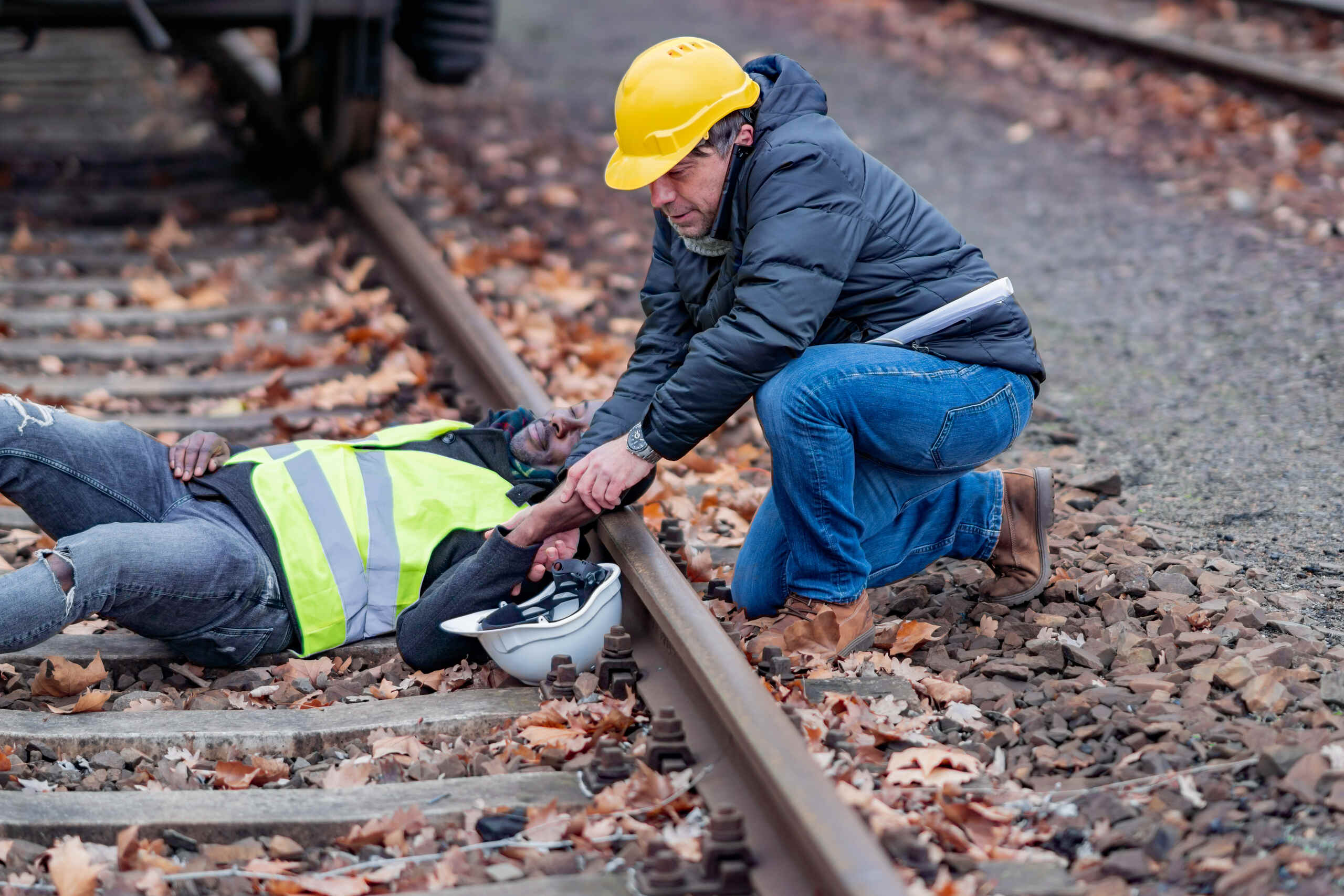 A man in a yellow hard hat kneels to assist an injured construction worker lying on railroad tracks after a workplace accident.