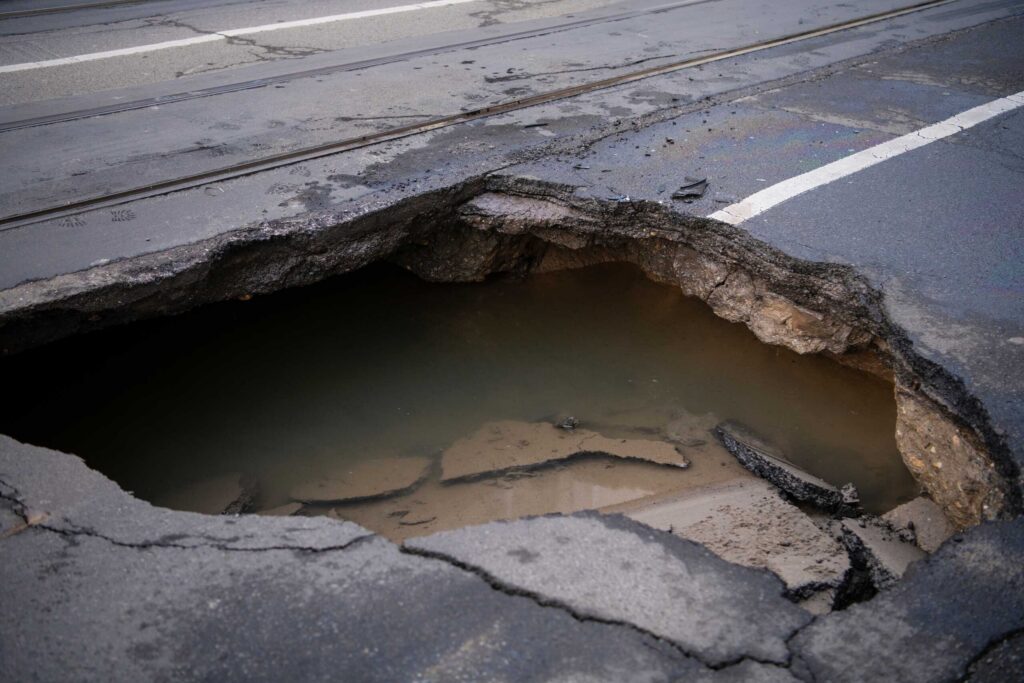 A large, deep pothole filled with murky water sits in the middle of a damaged asphalt road next to light rail tracks.