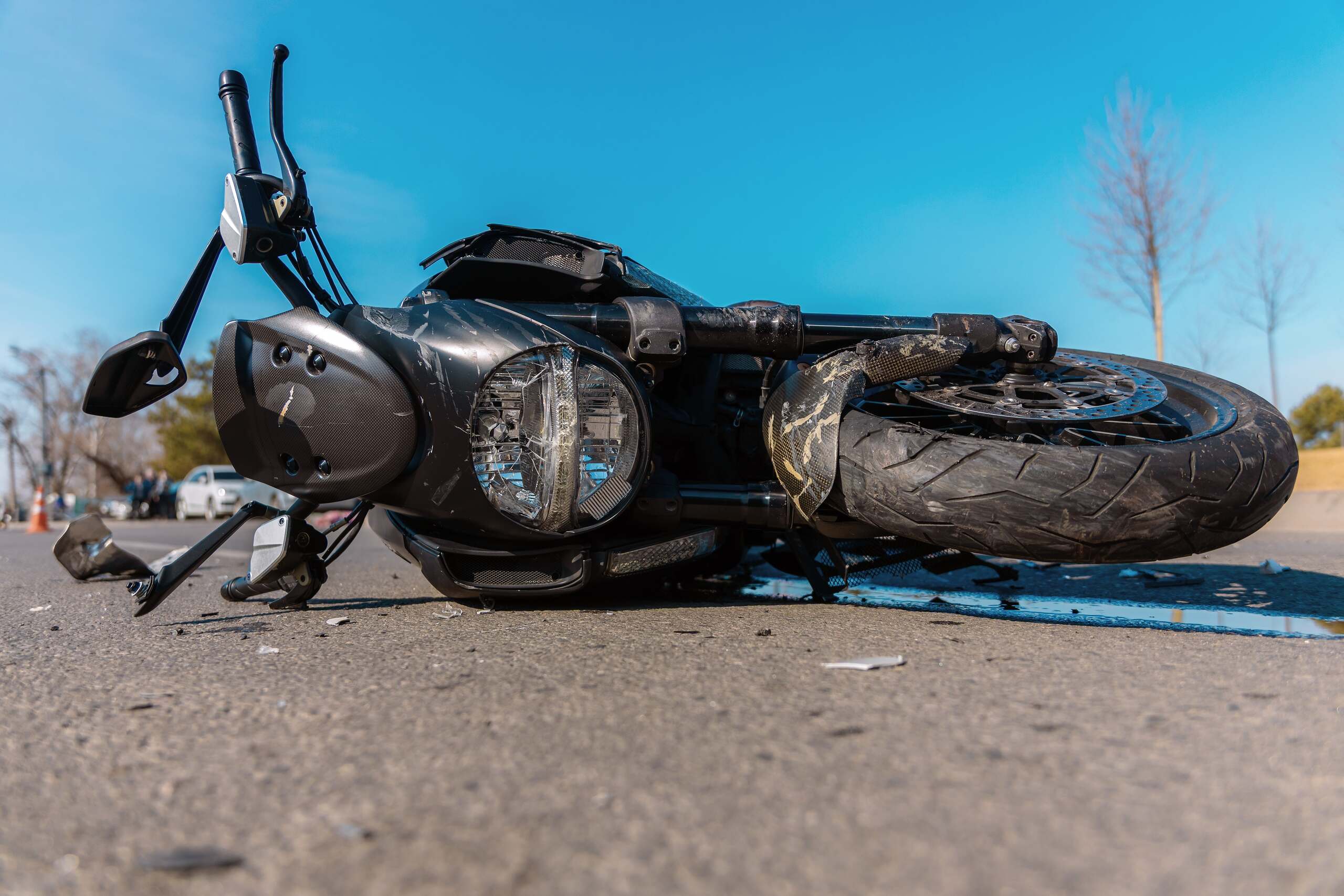 A wrecked black motorcycle lies on its side on a paved road with debris scattered nearby after an accident.