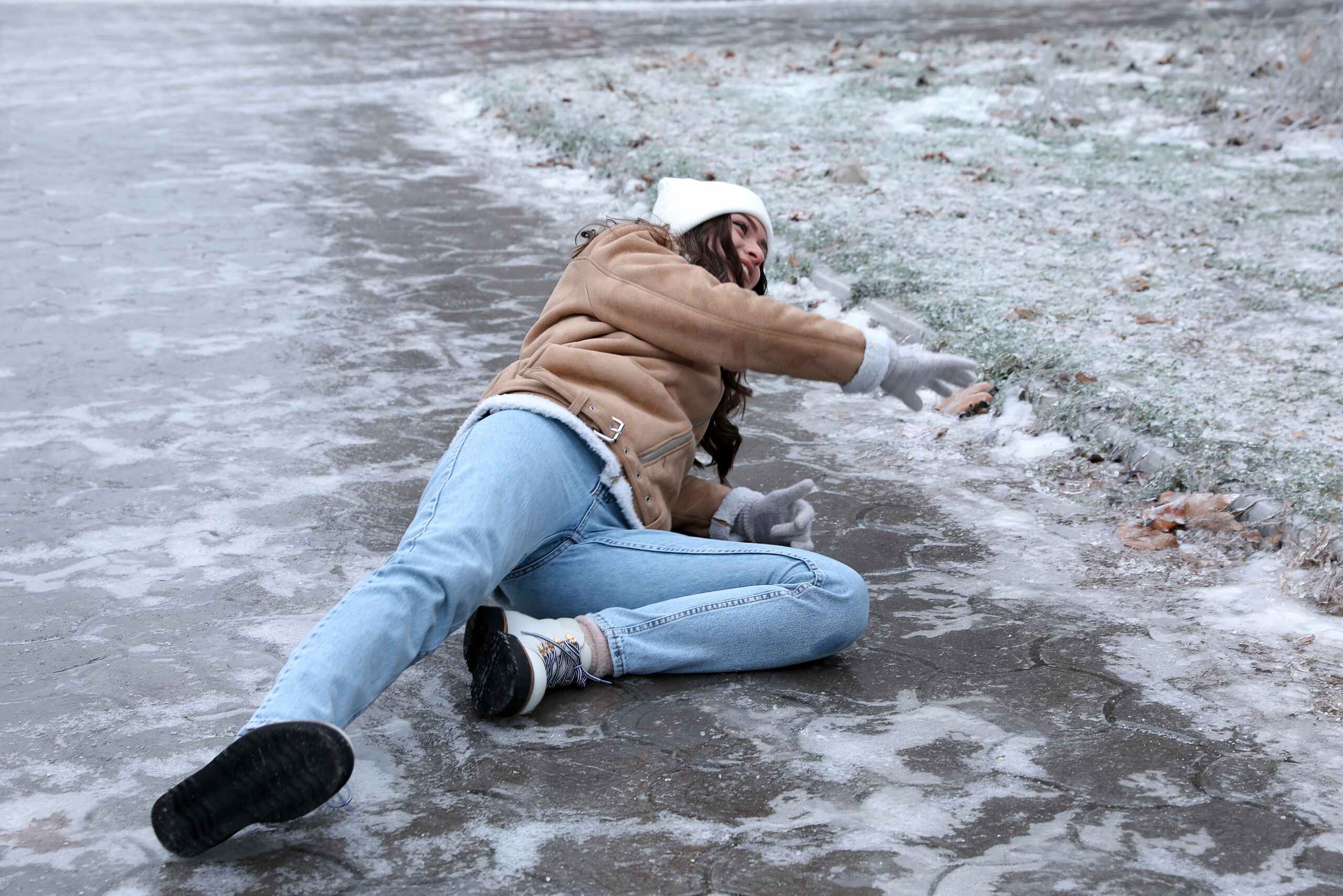 A woman in winter clothing slips and falls backward on an icy sidewalk, reaching out to break her fall.