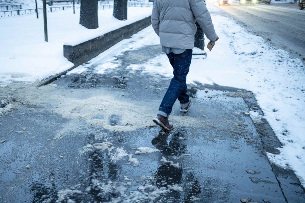 A person walks carefully across a slippery, wet sidewalk covered in patches of melting snow and ice near a busy road.