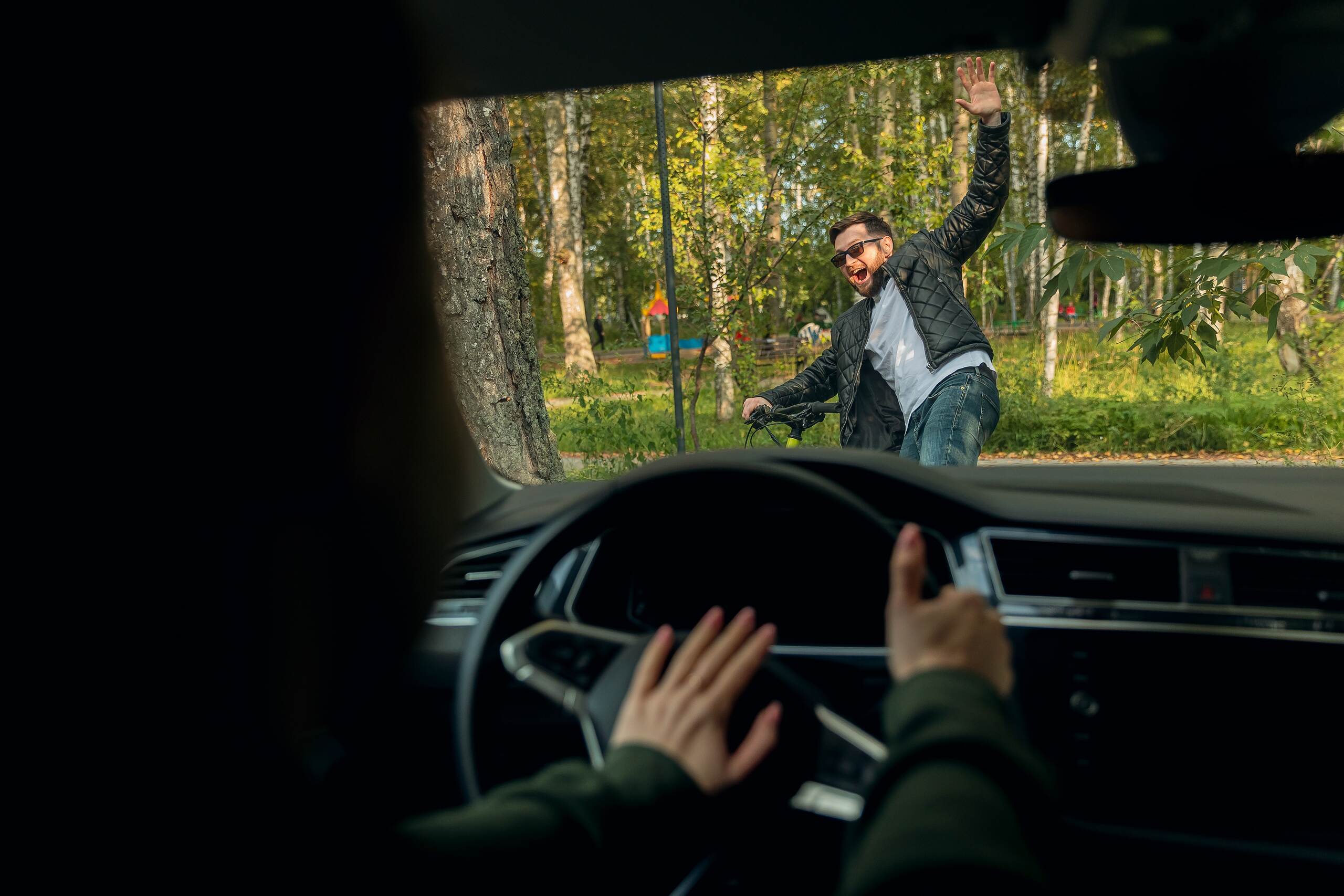 A view from inside a car looking through the windshield at a cyclist falling over after a near-collision or accident on a tree-lined road.