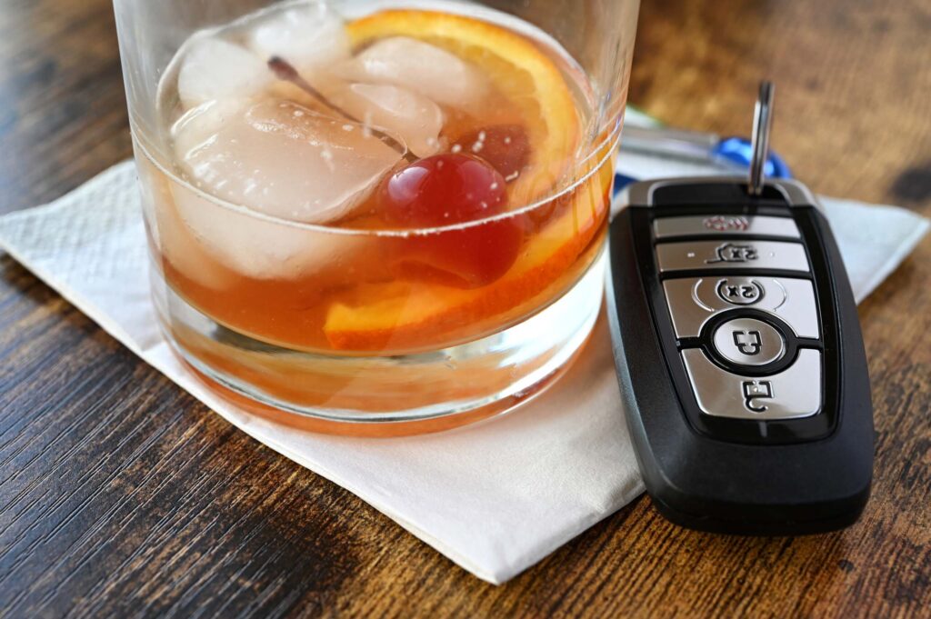 A close-up of a glass of amber-colored alcohol next to a set of car keys on a wooden table. Utah drunk driving accident lawyers advocate for victims to hold impaired drivers accountable for the physical and financial damages they cause.