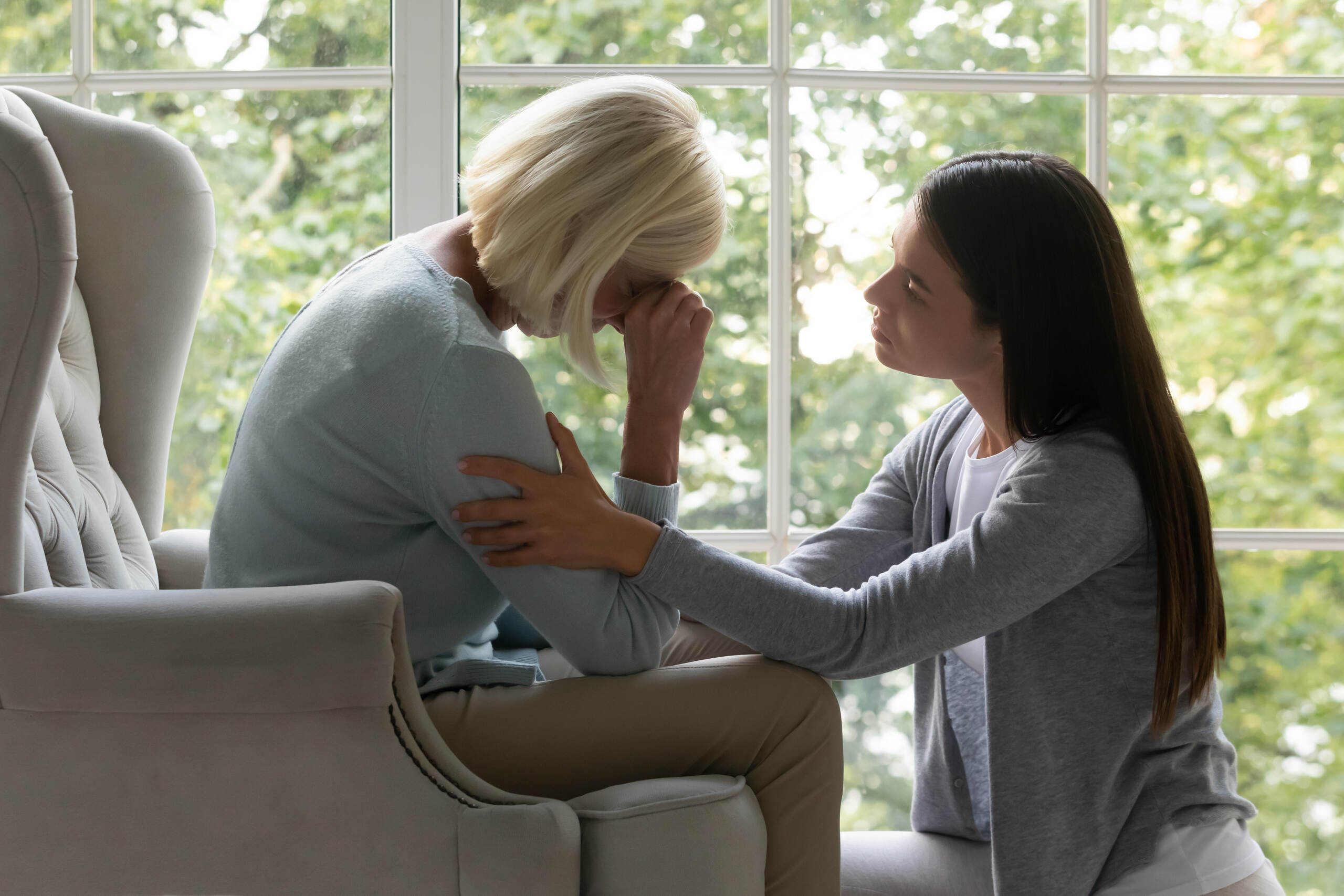 An indoor scene where a younger woman supportively holds the arm of an older, blonde woman who is seated and crying with her hand to her face.
